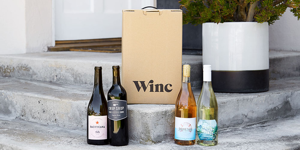 Four bottles of wine at a front door and a brown bag that says Winc. 