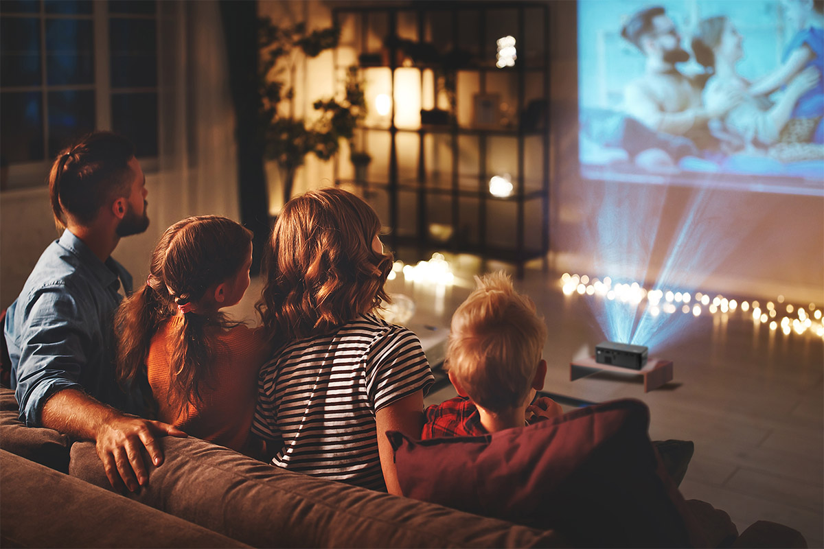 a young man, a girl in pigtails, a woman with short hair, and a young boy sit on a couch and look at family vacation photos projected onto their living room wall